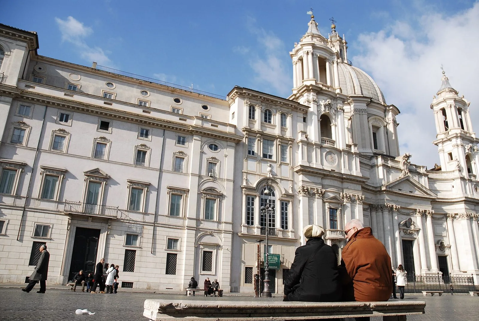 La chiesa barocca di Sant'Agnese in Agone e i palazzi chiari di Piazza Navona sotto un cielo azzurro, con persone sedute di spalle su una panchina di pietra