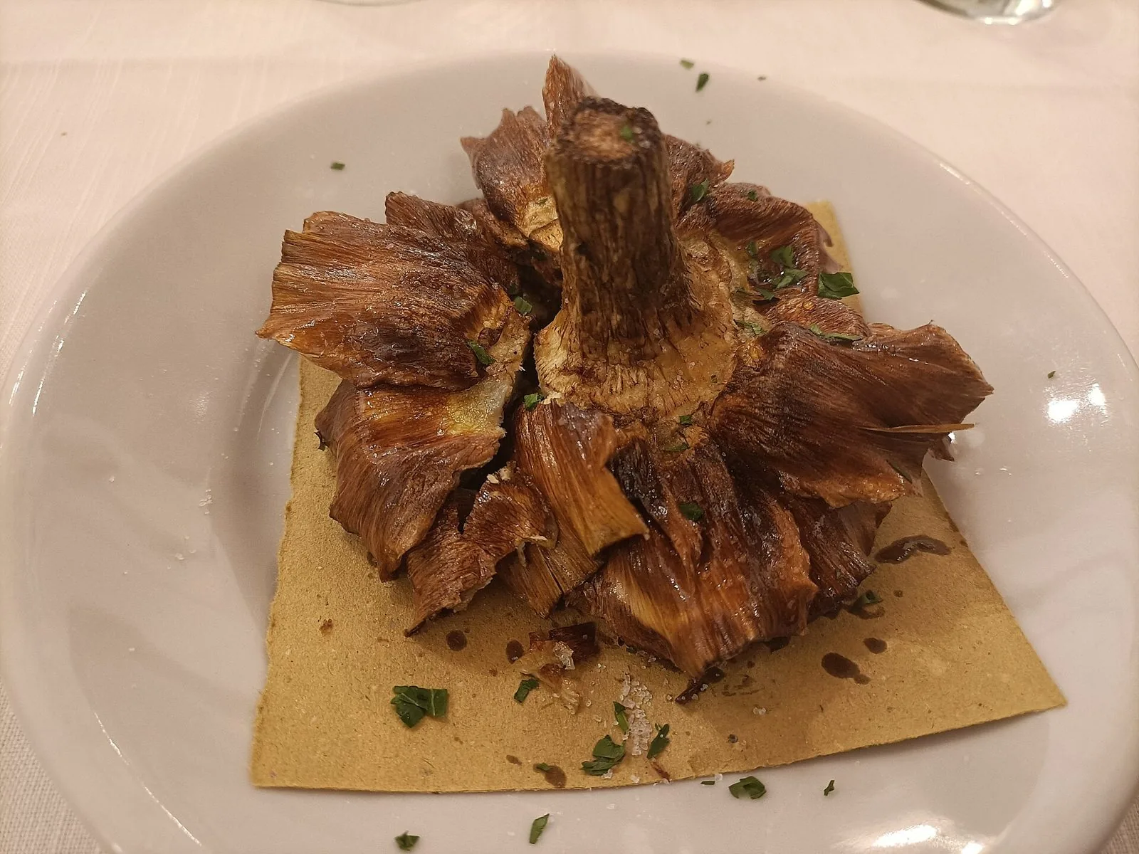 Un carciofo alla giudia fritto e dorato, con le foglie aperte a fiore, servito su un piatto bianco con prezzemolo tritato