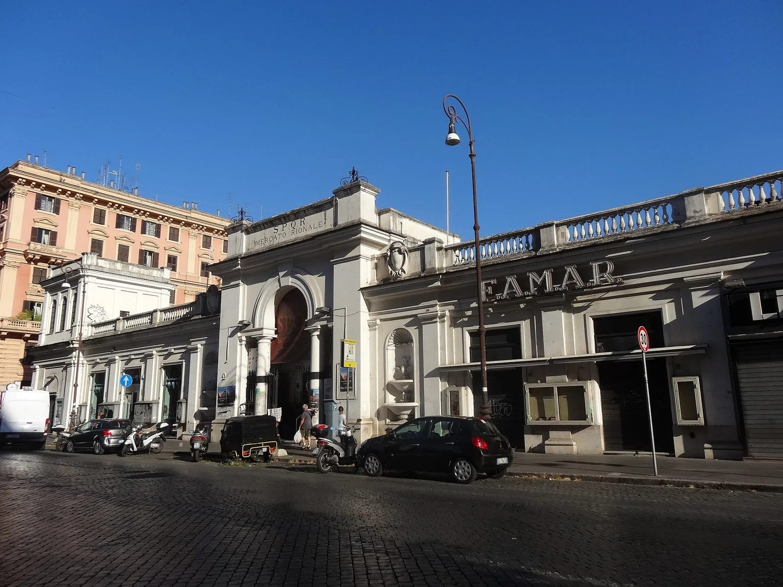 La facciata bianca neoclassica del mercato rionale di piazza dell'Unità in via Cola di Rienzo, con il portale ad arco e la scritta SPQR Mercato Rionale, sotto un cielo azzurro