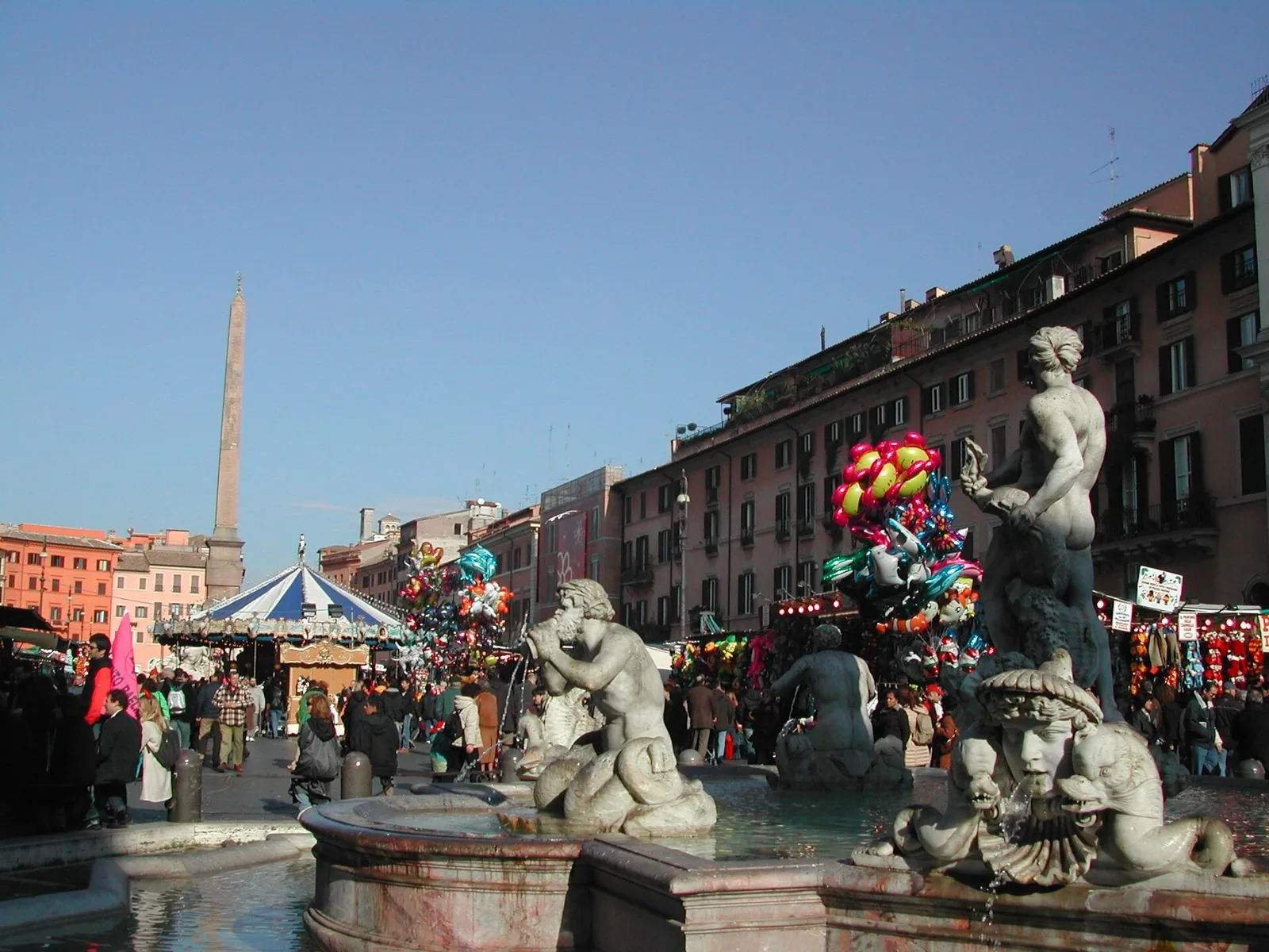 Piazza Navona durante il periodo natalizio, con la Fontana del Moro in primo piano, bancarelle, palloncini colorati, una giostra e l'obelisco sullo sfondo