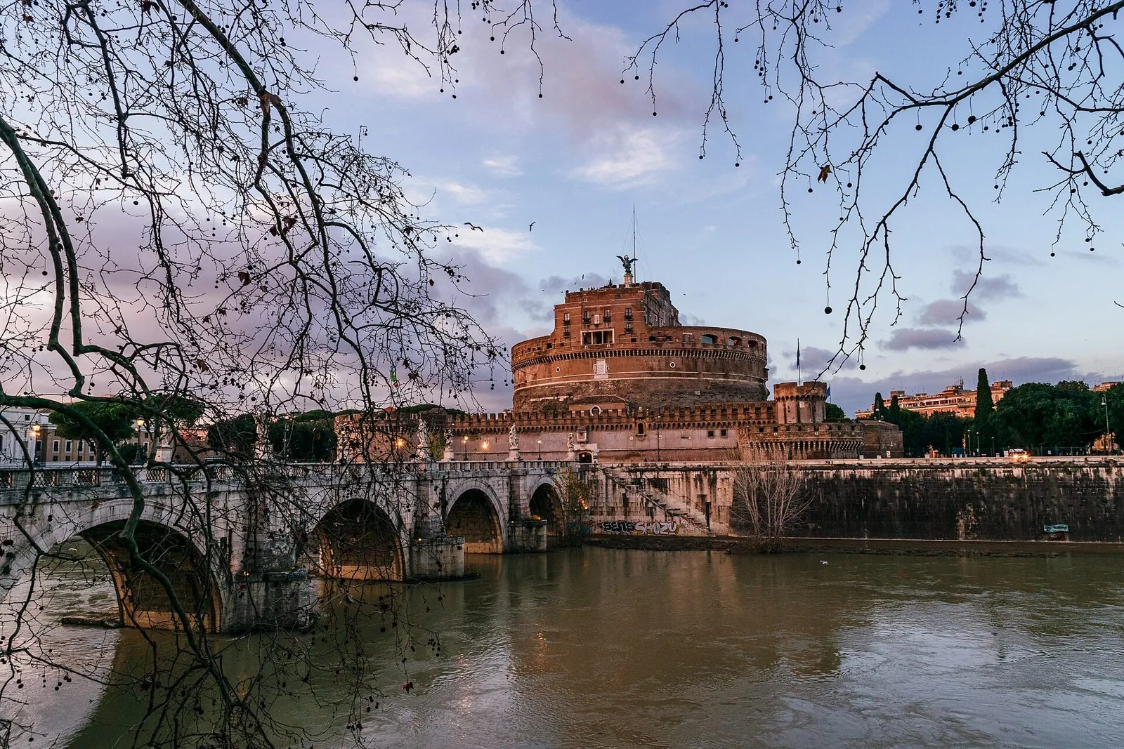Ponte Sant'Angelo con le sue arcate sul Tevere visto dal lungotevere tra i rami spogli degli alberi, con Castel Sant'Angelo e i muraglioni del fiume sullo sfondo al tramonto