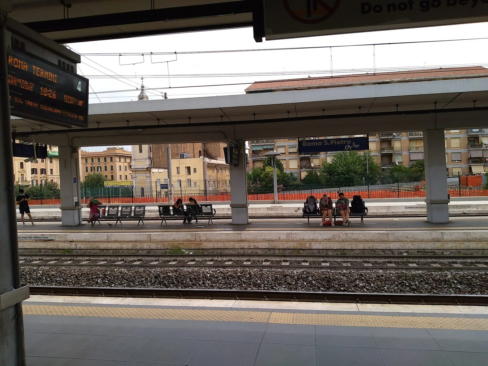 Banchine della stazione di Roma San Pietro con l'insegna blu della stazione, pensiline, binari e un tabellone con un treno per Roma Termini